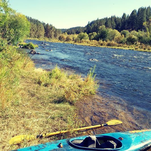 Old whitewater kayak on the bank of a burbling blue river with pine trees and willows